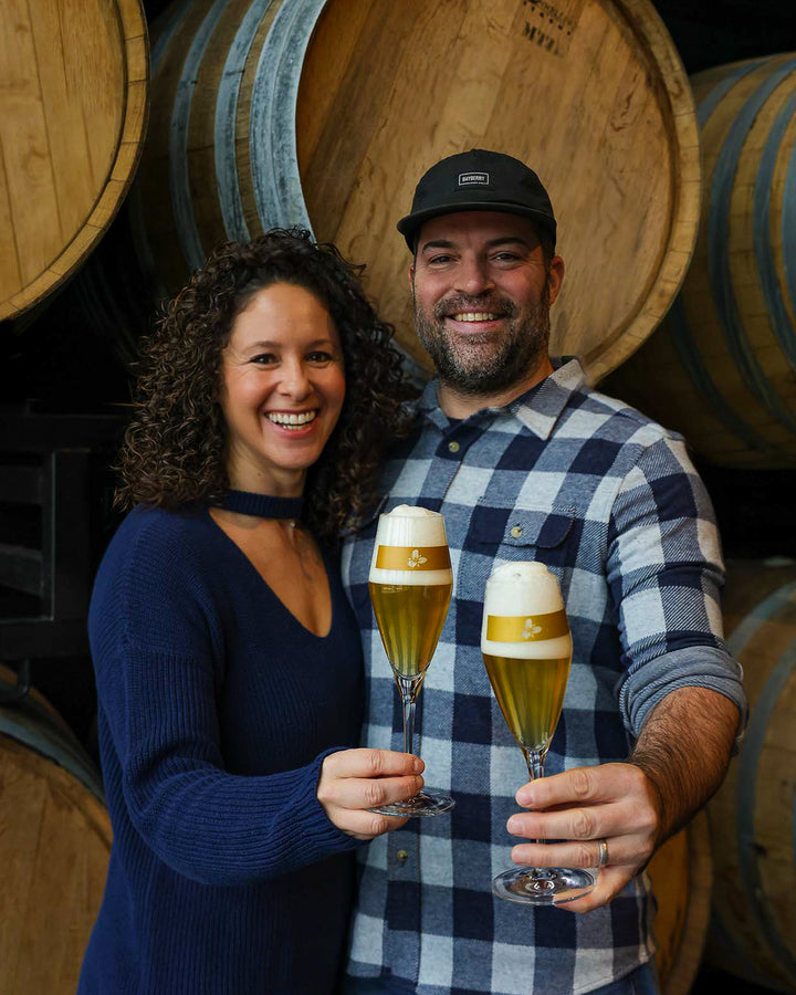 Esther and JC Tetreault cheersing with flute glasses in front of barrels
